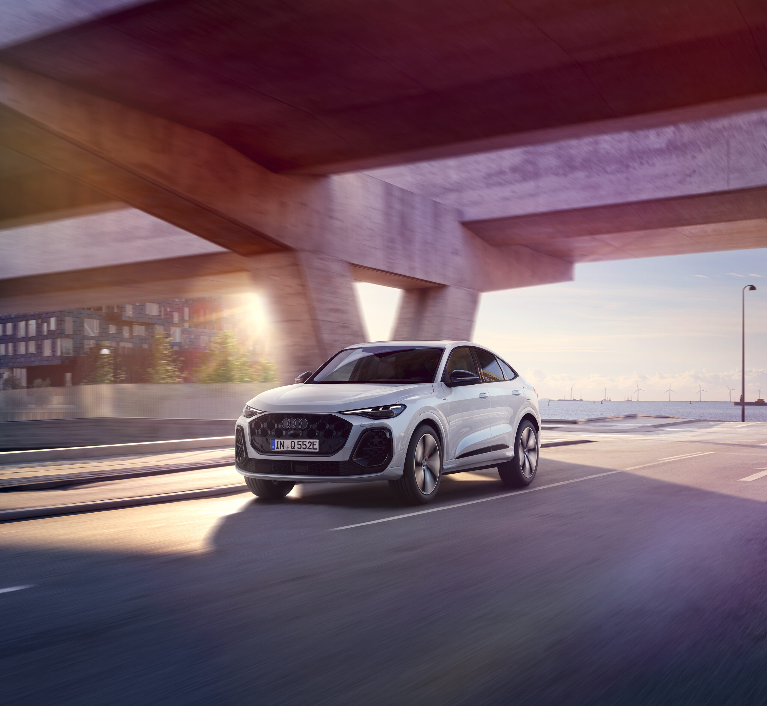 Ein weißer Audi Q5 Sportback fährt dynamisch unter einer modernen Betonbrücke entlang, während im Hintergrund die Morgensonne durchscheint und Windräder am Horizont sichtbar sind.