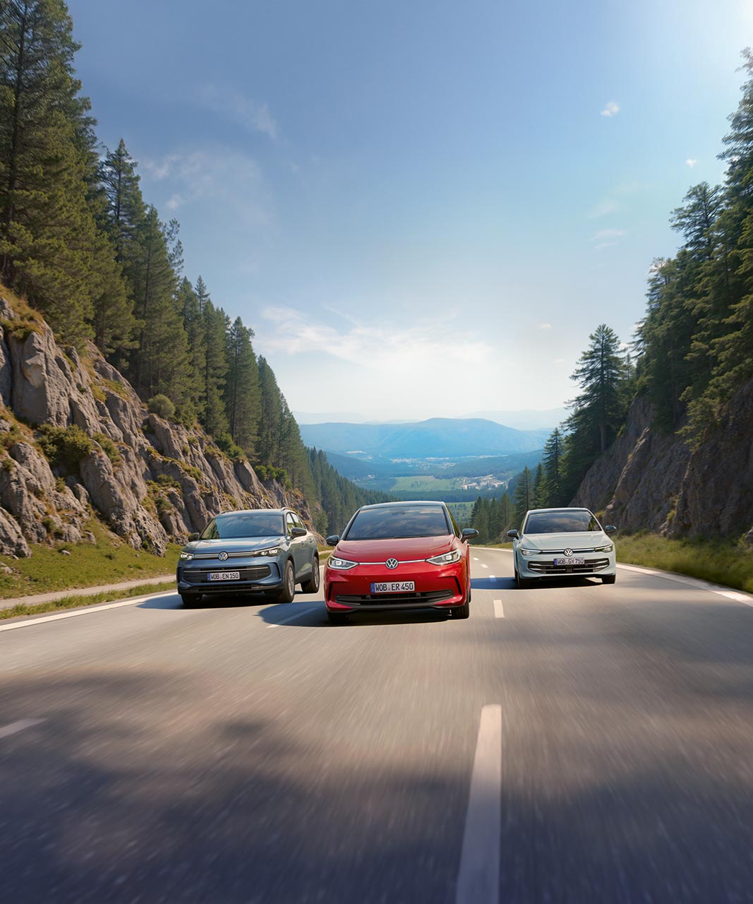 Drei Volkswagen-Modelle fahren nebeneinander auf einer Landstraße durch eine von hohen Felswänden und dichten Nadelwäldern gesäumte Berglandschaft, mit Blick auf ein weites Tal und ferne Gebirgszüge unter blauem Himmel